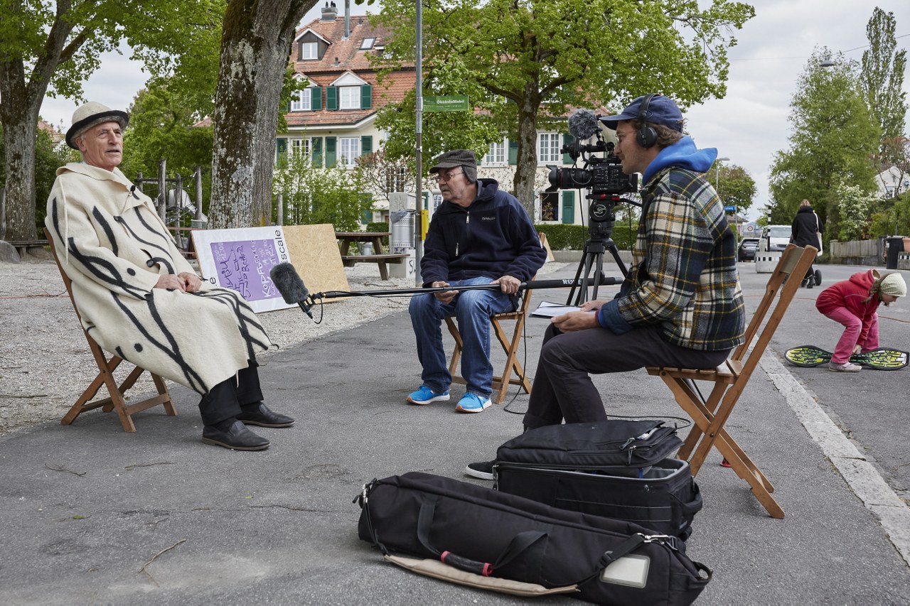 Das Bild zeigt einen älteren Herren im Mantel auf einem Klappstuhl auf einem öffentlichen Platz, der in ein Mikrofon erzählt.