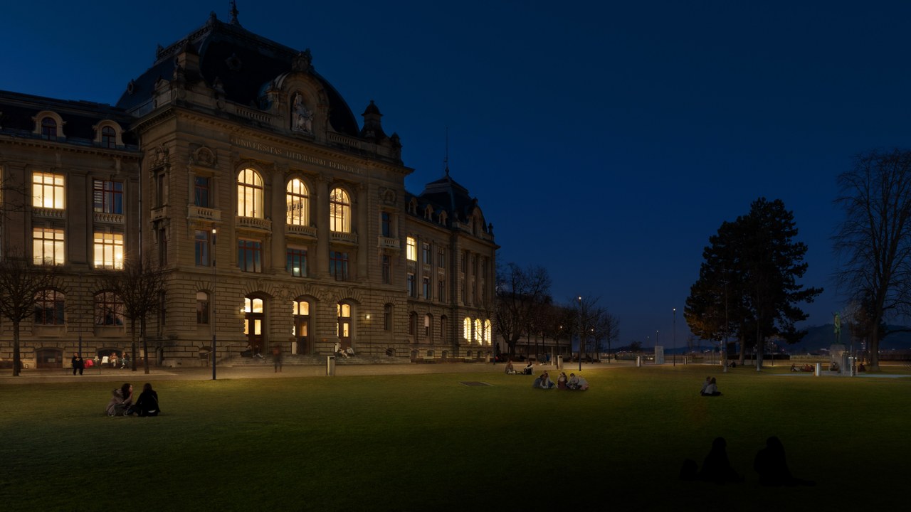 Das Bild zeigt das Hauptgebäude der Uni Bern bei Nacht, die Wiese mit sitzenden Menschen ist sanft beleuchtet.