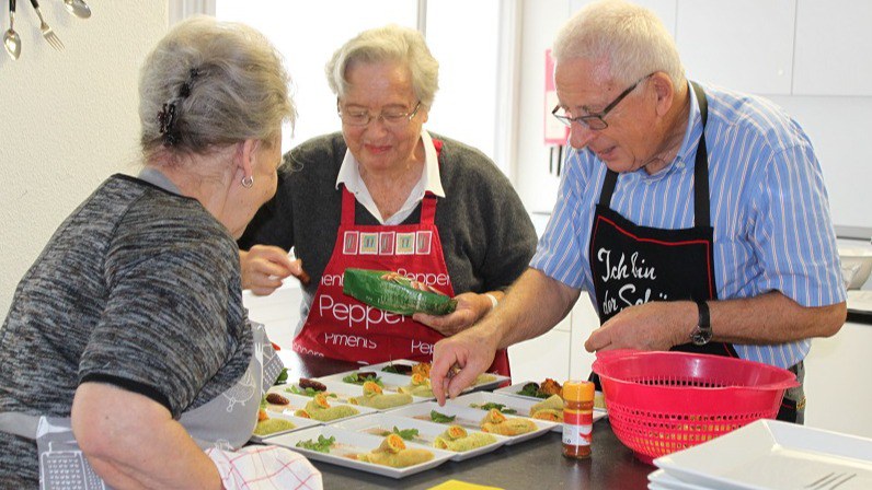 drei ältere Personen stehen gemeinsam um eine Kochinsel und tragen Küchenschürzen
