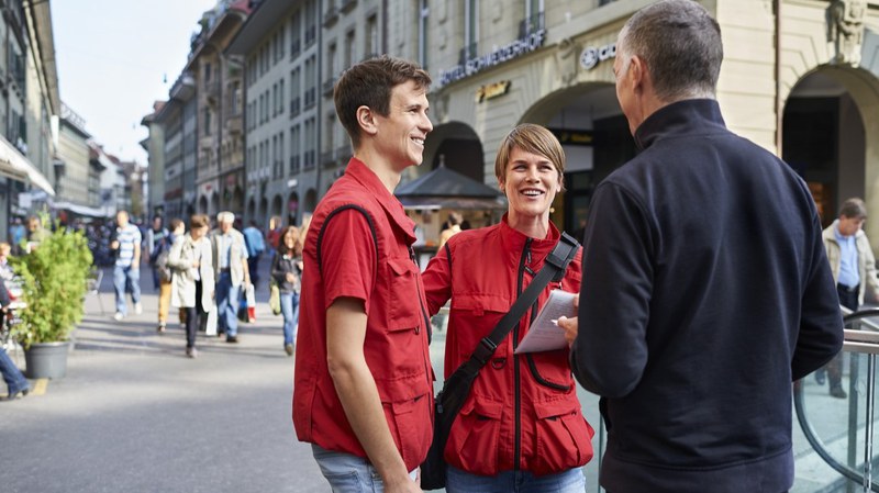 Zwei Personen mit roten Westen sprechen auf der Strasse mit einer dritten Person