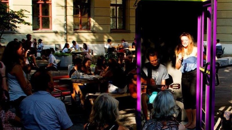 Zwei Personen treten mit Gitarre und Gesang in einer Telefonkabine auf. Darum herum sitzt Publikum.