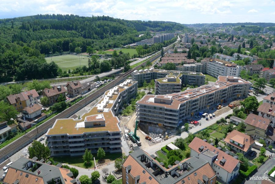 Das Bild zeigt eine Drohnenaufnahme der abgeschlossenen städtischen Überbauung Stöckacker Süd. Im Hintergrund ist der grüne Könzbergwald und dazwischen die Eisenbahnlinie Bern-Freiburg zu sehen.