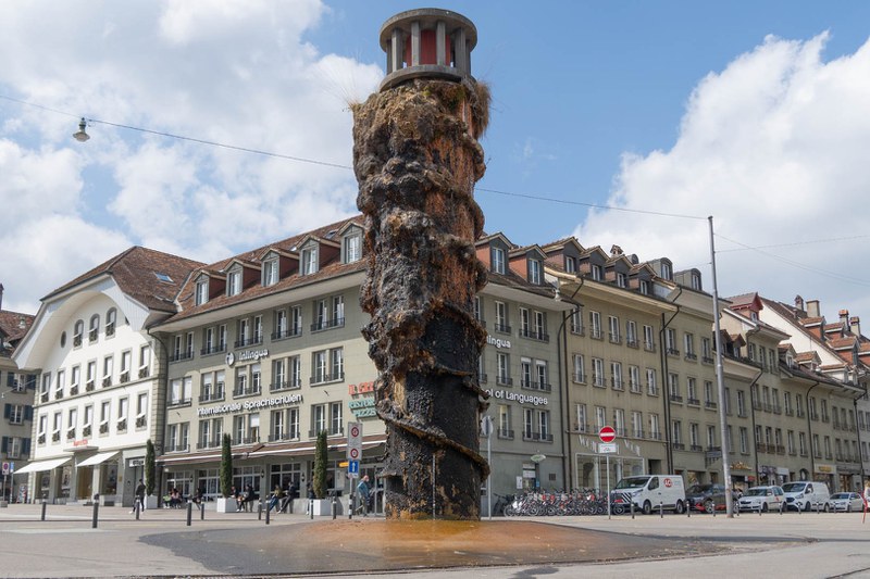 Das Bild zeigt den Meret-Oppenheim-Brunnen. Der 1983 eingeweihte Brunnen ist stark von Moospolster, Pflanzen und Gräser überwuchert, die - wie von der Künstlerin beabsichtigt - entlang der spiralförmig angelegten Wasserführung gewachsen sind.