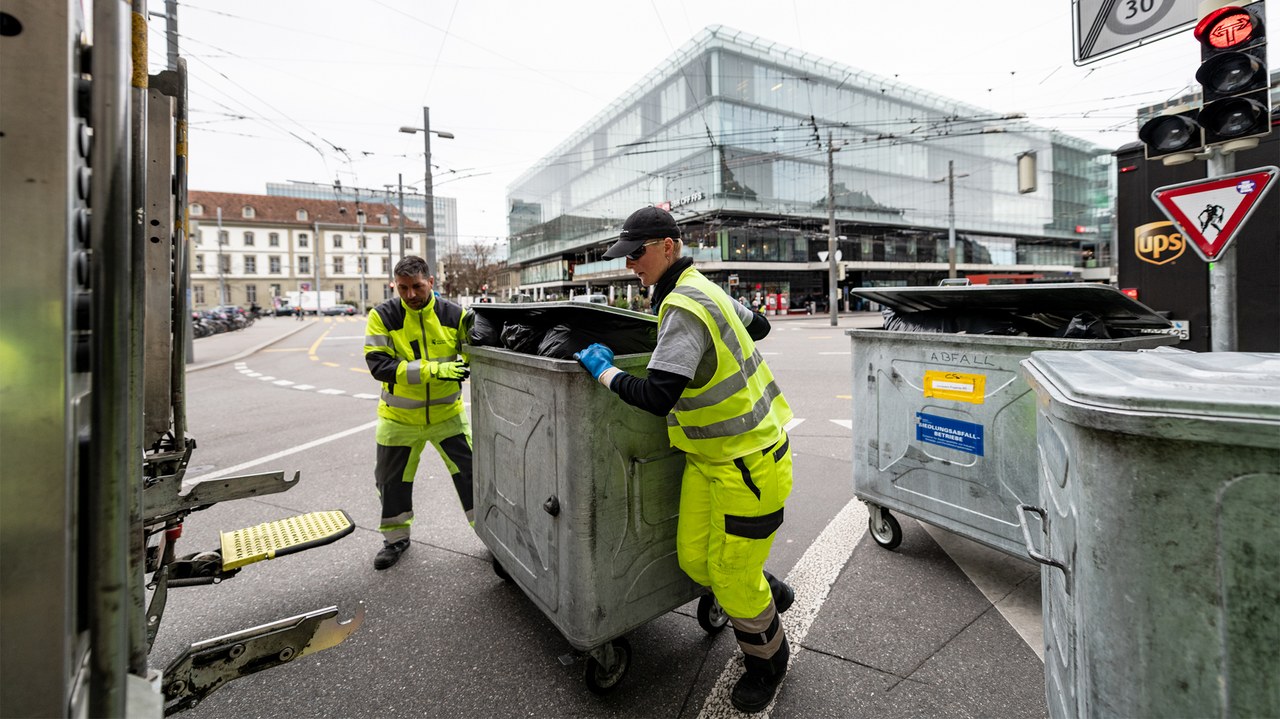 Zwei Mitarbeitende schieben einen Container mit schwarzen Säcken darin zum Kehrichtlastwagen.