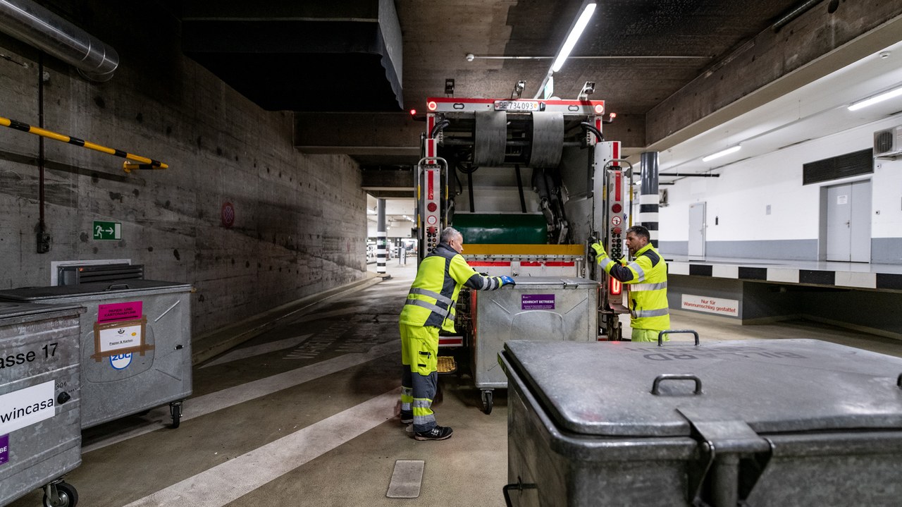 Es hat verschiedene Metallcontainer auf dem Bild. Einen davon sind zwei Mitarbeitende beim Kehrichtlastwagen am leeren.