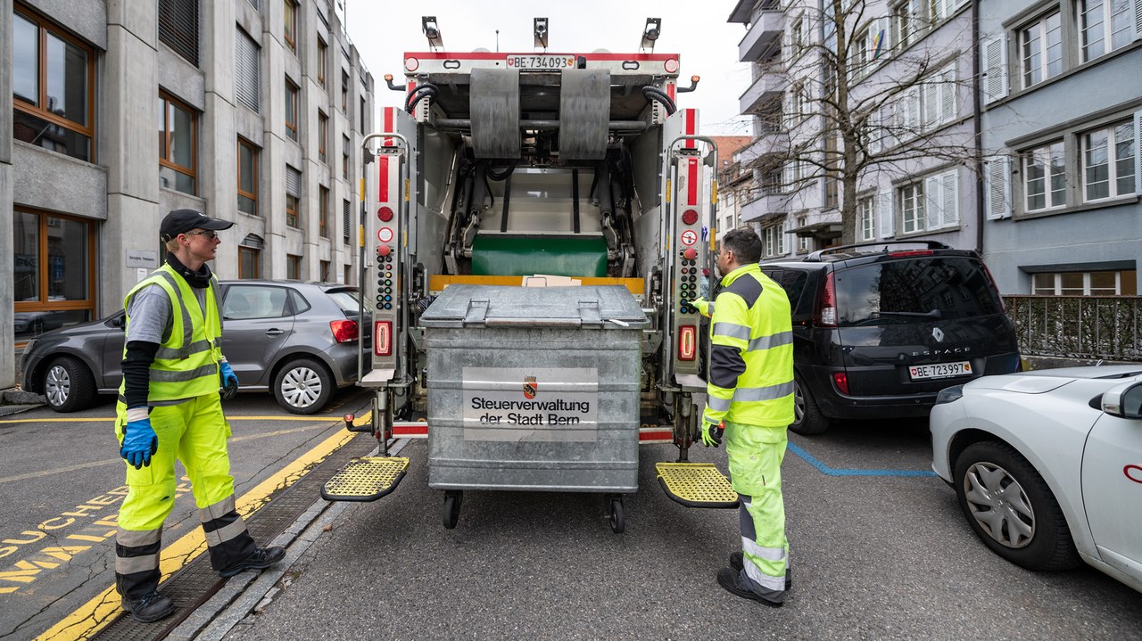 Zwei Mitarbeitende schauen zu, wie ein Container vom Kehrichtlastwagen geleert wird.