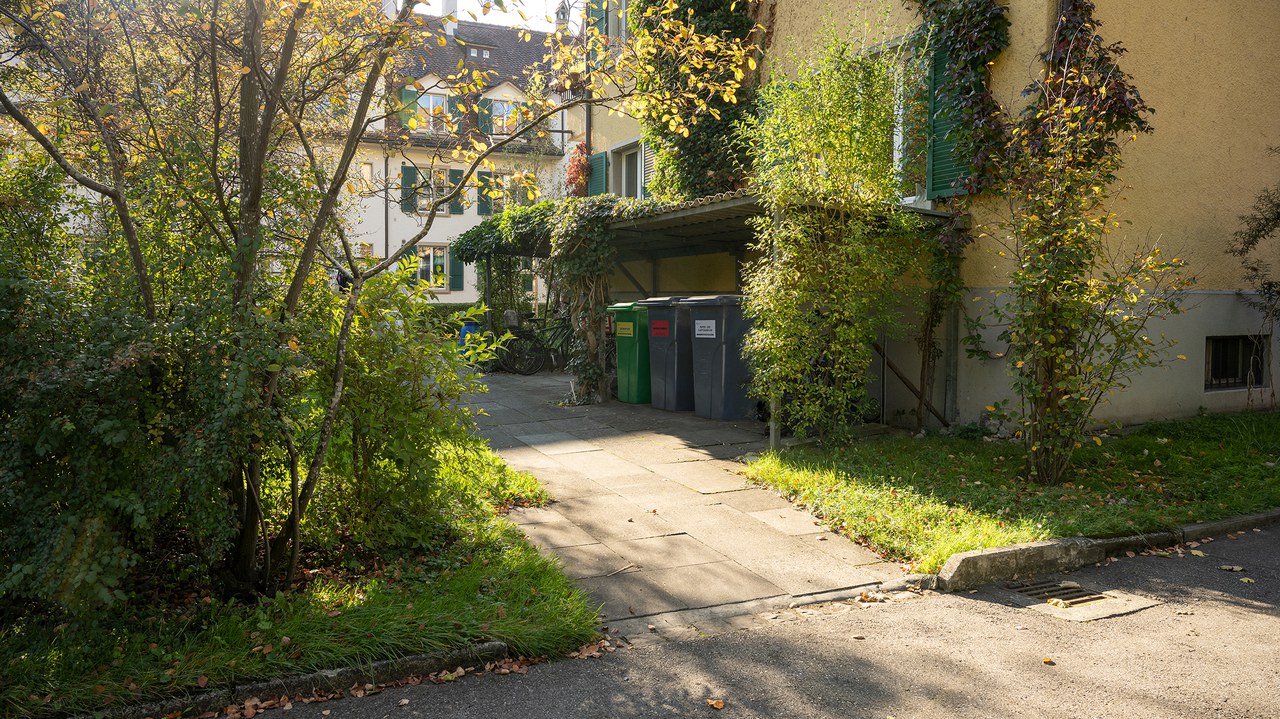 Der Zugang zu einem Haus, Plattenboden. Im Hintergrund stehen drei Container in einem Unterstand an der Hausfassade.