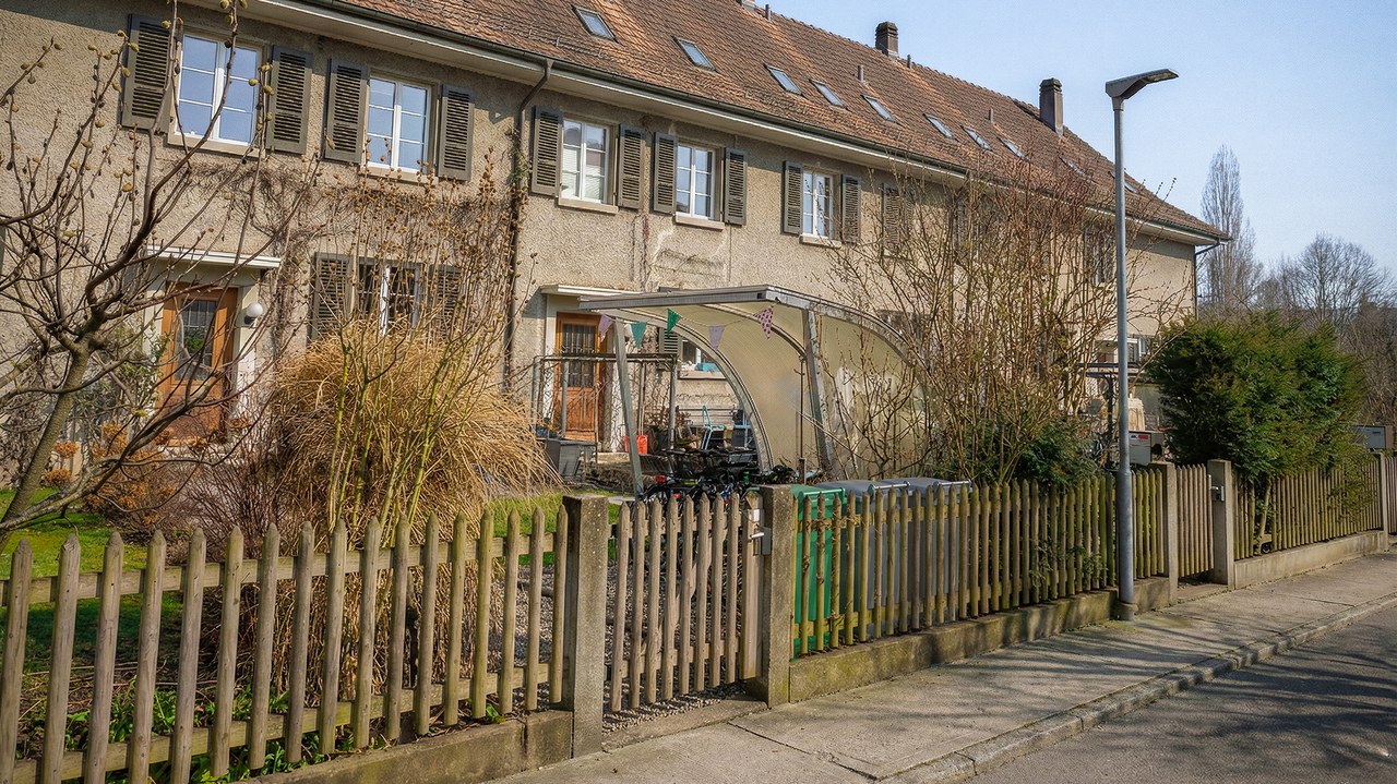 Reiheneinfamilienhäuser mit dem Vorgarten und der Strasse. Im Vorgarten stehen halb versteckt hinter einem Zaun Container.