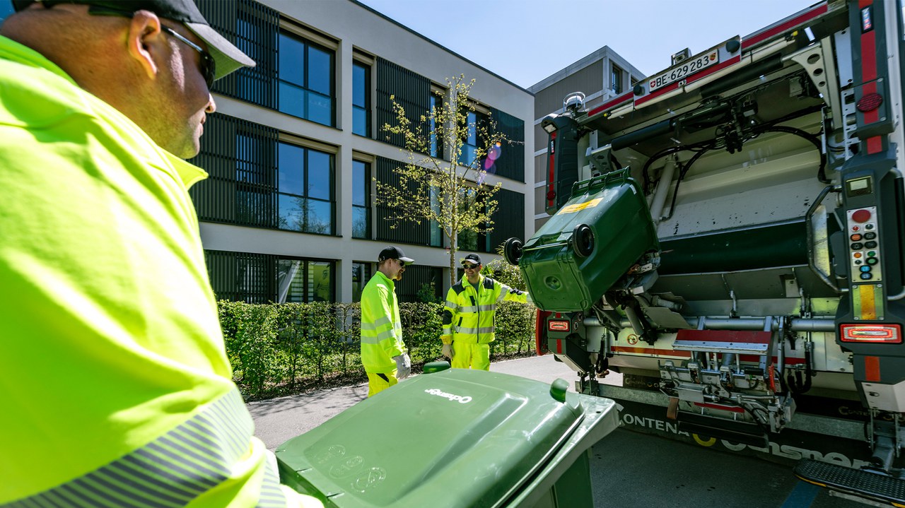 Mitarbeiter schiebt Grüncontainer zum Kehrichtwagen, wo bereits zwei andere Mitarbeitende einen Grüngut-Container am leeren sind.
