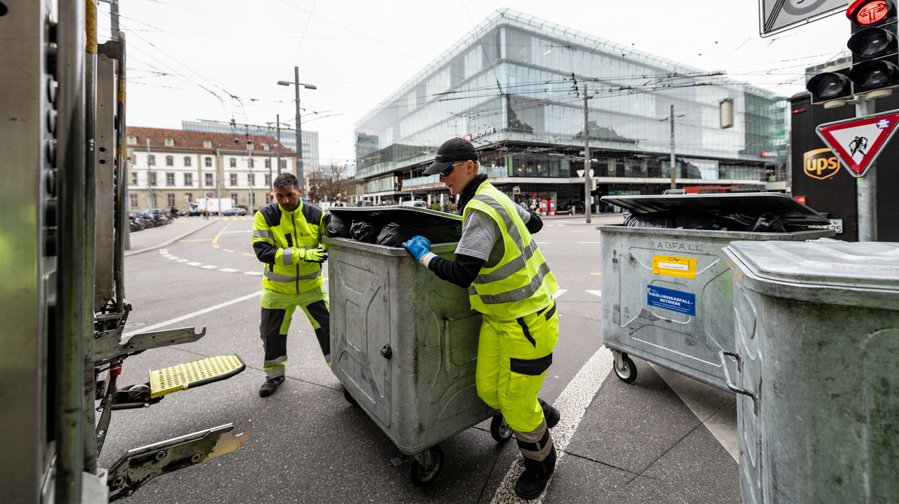 Zwei Mitarbeitende schieben einen Container aus Metall zum Kehrichtwagen.