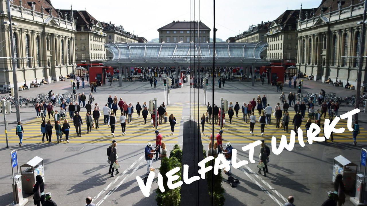 iele Menschen überqueren den Zebrastreifen beim Bahnhof Bern, Spiegelung an einer Glasfassade verstärkt die Szene. Text im Bild: „Vielfalt wirkt“.