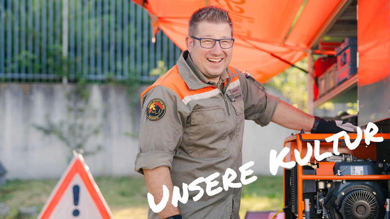 Lachender Zivilschutzmitarbeiter neben einem Notstromaggregat im Einsatz. Im Bild ist der Schriftzug "unsere Kutur" enthalten.