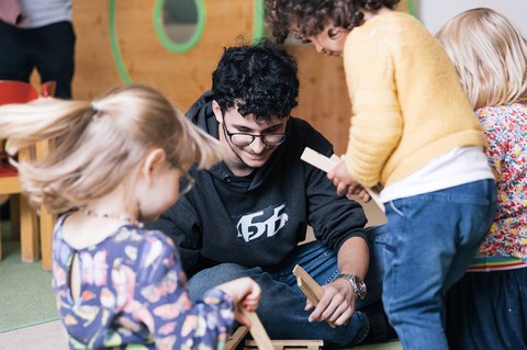 Junger Mann sitzt am Boden einer Kita und spielt lächelnd mit drei Kindern, die Holzklötze in der Hand halten.