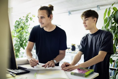 Zwei junge Männer arbeiten konzentriert gemeinsam an einem Schreibtisch vor einem Bildschirm