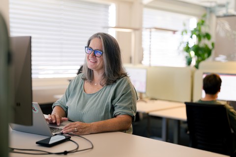Frau arbeitet lächelnd am Laptop in einem Grossraumbüro.