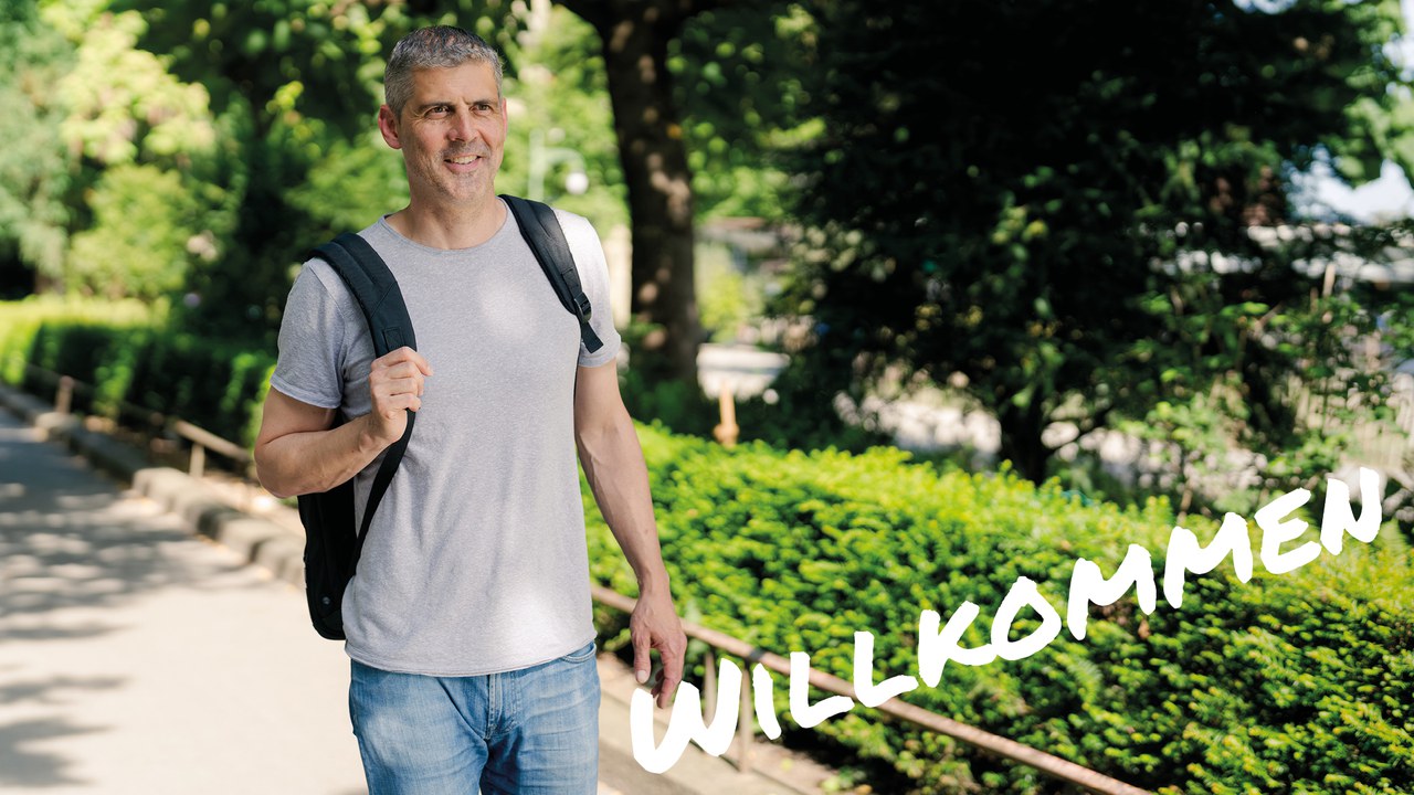 Mann geht lächelnd auf einem sonnigen Weg im Grünen. Text im Bild: „Willkommen“