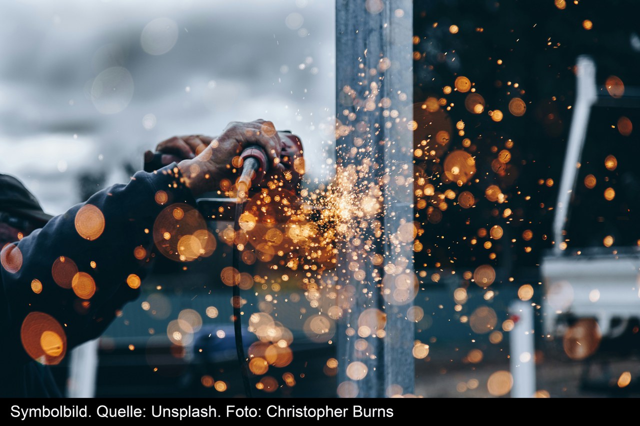 Allgemeines Industriebild. Zwei Hände die eine Stahlstange halten. Im Vordergrund verschwommen Punkte von heisser Glut.