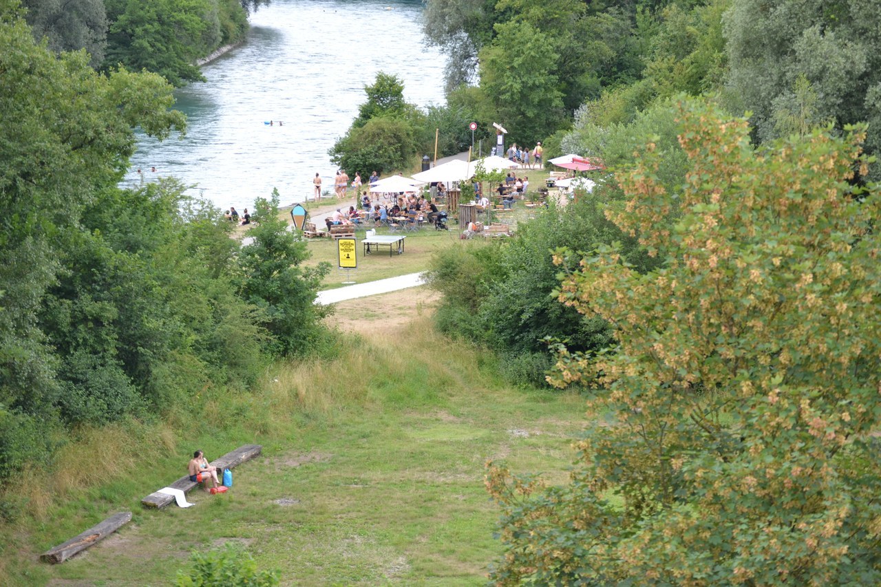 Das Foto zeigt viele Bäume, Grünflächen und die Aare mit Schwimmenden. Gut sichtbar ist die Aarebar mit vielen Gästen, die als Pop-up während den Sommermonaten das Areal belebt.