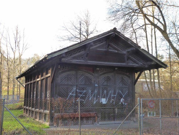 Das Foto zeigt den alten Schopf aus Holz, umgeben von einem Maschendrahtzaun. Die Fassade des Schopfs ist unleserlich verschmiert.