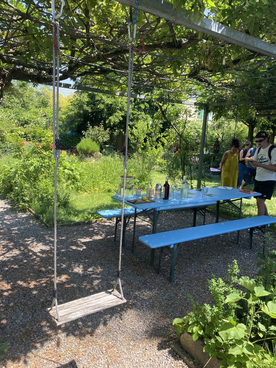 Schaukel im Vordergrund, blauer Gartentisch und Bank im Mittelgrund und im Hintergrund sind Menschen am Sprechen und Essen zu sehen. Die Umgebung ist sehr grün und befindet sich in einem Privatgarten